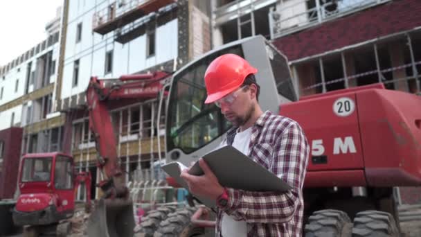 Кавказский инженер-архитектор держит папку с бумагами на фоновом экскаваторе, строительную технику на стройплощадке. Строитель просматривает бизнес-план, архитектурные документы — стоковое видео