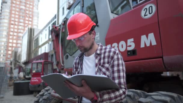 Mladý stavební manažer zkontroluje technickou dokumentaci na staveništi poblíž těžkého traktoru nebo bagru. Kontrolní zpráva Foremana — Stock video