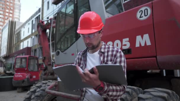 若い建設管理者は、重機トラクターや掘削機の近くの建設現場にいる間、技術文書をチェックします。陪審員長の報告書 — ストック動画
