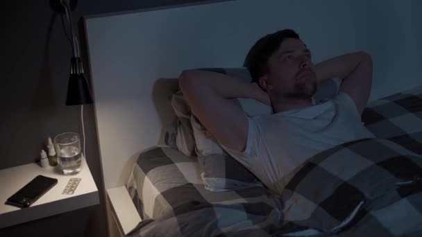 Fiatalember nézi a plafont éjszaka, miközben fekszik az ágyban, alvás helyett, ellenőrzi az időt a mobilján, álmatlanságban szenved. Elégedetlen férfi fekszik az ágyban képtelen elaludni — Stock videók