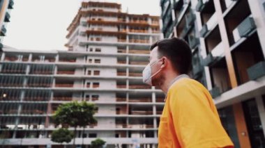 Professional builder in safety equipment KN 95 mask and hard hat at construction site talks to employee, shows hand to project. Masked foreman in orange uniform points to house under construction