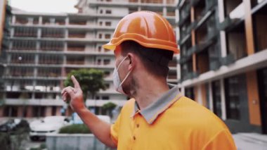 Construction worker in protective helmet, mask and orange uniform is discussing details of project at construction site looking into camera and showing with his hand at building under construction