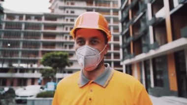 Professional builder in safety equipment KN 95 mask and hard hat at construction site talks to employee, shows hand to project. Masked foreman in orange uniform points to house under construction