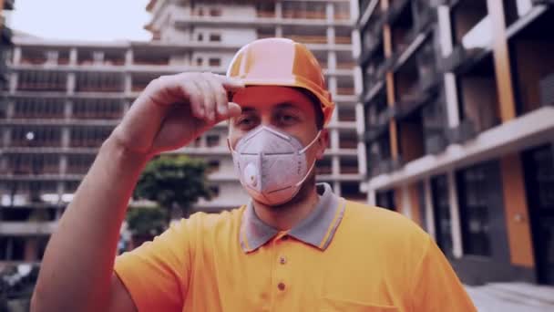 Foreman i skyddshjälm och respirator kontrollerar byggprocessen och talar med en arbetare tittar på kameran på en byggarbetsplats mot bakgrund av en byggnad — Stockvideo