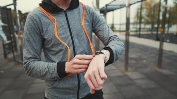 Man measuring pulse on sports watch during training jumping to ropes outdoors. Sporty male with orange rope around his neck setting up smart watch. Records the results of workout exercises on device — Stok video