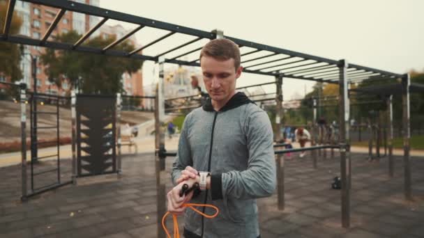 Young sportsman checking heart rate on sports watch during exercise jumping on rolling pin at street gym. Sporty man checking pulse after exercising with jumping rope. Checking activity wrist tracker — Vídeo de Stock