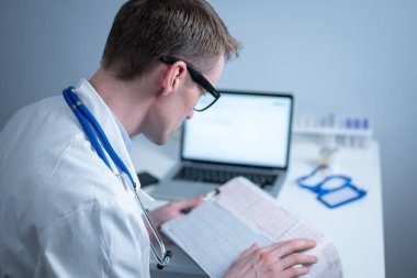 Gözlüklü ve beyaz laboratuvar önlüklü yakışıklı doktor hasta kalbinin hastane ofisinde elektrokardiyografik muayenesinin sonuçlarını inceliyor. Kardiyolog muayenehanede kardiyograma bakıyor..