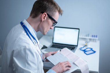 Gözlüklü ve beyaz laboratuvar önlüklü yakışıklı doktor hasta kalbinin hastane ofisinde elektrokardiyografik muayenesinin sonuçlarını inceliyor. Kardiyolog muayenehanede kardiyograma bakıyor..