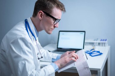 Gözlüklü ve beyaz laboratuvar önlüklü yakışıklı doktor hasta kalbinin hastane ofisinde elektrokardiyografik muayenesinin sonuçlarını inceliyor. Kardiyolog muayenehanede kardiyograma bakıyor..