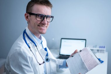 Gözlüklü ve beyaz laboratuvar önlüklü yakışıklı doktor hasta kalbinin hastane ofisinde elektrokardiyografik muayenesinin sonuçlarını inceliyor. Kardiyolog muayenehanede kardiyograma bakıyor..