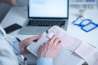 Kardiyolog EKG çıktısı okuyor. Doktor elektrokardiyogramı analiz ediyor. Pratisyen hasta test sonuçlarını inceliyor. Tıbbi ve sağlık hizmetleri konsepti. Hekim muayenehanesinde kardiyograma bakıyor..