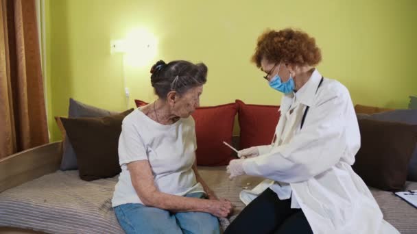 Una mujer mayor está siendo examinada para el coronavirus en casa. Un médico con bata de laboratorio toma hisopo de covid 19 de la mujer mayor de la garganta en el asilo de ancianos. Prueba rápida del antígeno durante la pandemia del coronavirus — Vídeo de stock