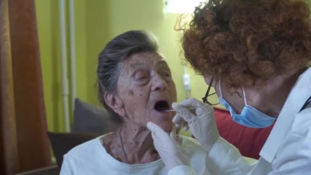 一名老年妇女正在家中接受验尸检查。一名穿着实验室外套的医生在疗养院从老年女性喉咙处接过肠子19号。大肠病毒大流行期间的快速抗原检测 — 图库视频影像