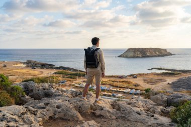 Man with backpack is hiking along a rocky coast at sunset near the Mediterranean sea in Cyprus. Male tourist admires the view Yeronisos, or Holy Island, lies off the coast of Agios Georgios Pegeias