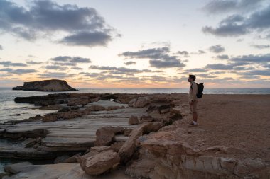 Sea sunset view. Man with backpack on rocks with beautiful view of Yeronisos Island near coast of Agios Georgios Pegeias. Guy enjoying ocean horizon, panoramic sunset in cyprus on mediterranean sea