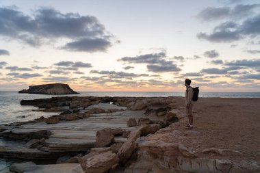 Male tourist with backpack watching beautiful view of Yeronisos Holy Island near coast Agios Georgios Pegeias In Cyprus on sunset. Man hiker looks into distance at deserted island in mediterranean sea