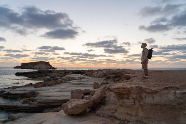 Man with backpack is hiking along a rocky coast at sunset near the Mediterranean sea in Cyprus. Male tourist admires the view Yeronisos, or Holy Island, lies off the coast of Agios Georgios Pegeias
