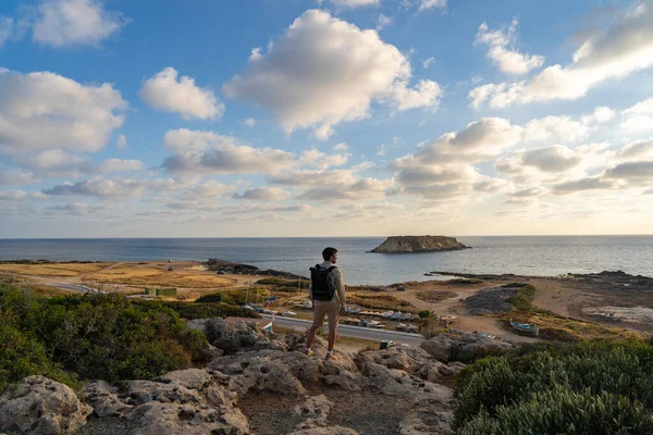 Male tourist with backpack watching beautiful view of Yeronisos Holy Island near coast Agios Georgios Pegeias In Cyprus on sunset. Man hiker looks into distance at deserted island in mediterranean sea — ストック写真