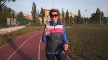 Elderly woman with Nordic walking poles on treadmill, rubber stadium stopped to quench her thirst, drink water from clear bottle. Athletic senior woman. Active retirement, healthy lifestyle concept