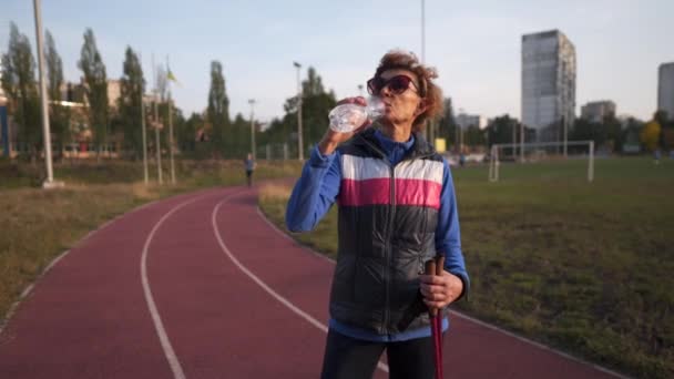 Happy senior woman drinks water from plastic bottle while training Nordic walking in city stadium. Tired elderly female refreshing herself after physical activity outdoors with Nordic walking sticks — Wideo stockowe