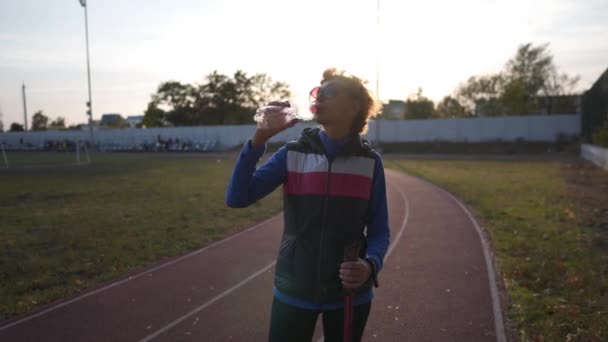 Gelukkig senior vrouw drinkt water uit plastic fles tijdens het trainen Nordic walking in het stadion van de stad. Moe oudere vrouw verfrissend na lichaamsbeweging buiten met Nordic walking sticks — Stockvideo