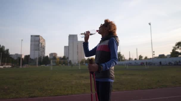 Water balance, importance drinking water during exercise. Middle age woman resting after Nordic walking and drinking water at stadium. Elderly female with walking sticks quenches thirst field workout — ストック動画