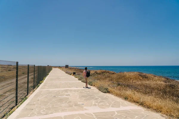 Man tourist with backpack walk along walking path at rocky coast of Mediterranean Sea in city Paphos of Cyprus in summer sunny hot weather. Coastal Broadwalk. Explore island of Cyprus on foot. Hiking — Stock Photo, Image