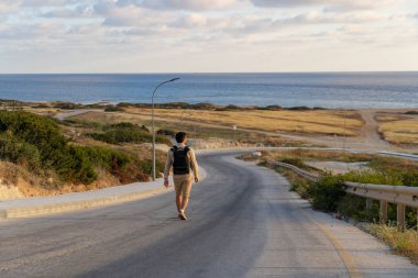Man tourist with backpack walks down mountain along asphalt road towards mediterranean sea in cyprus in area of Agios Georgios Pegeias. Rear view of backpacker walking along highway to ocean