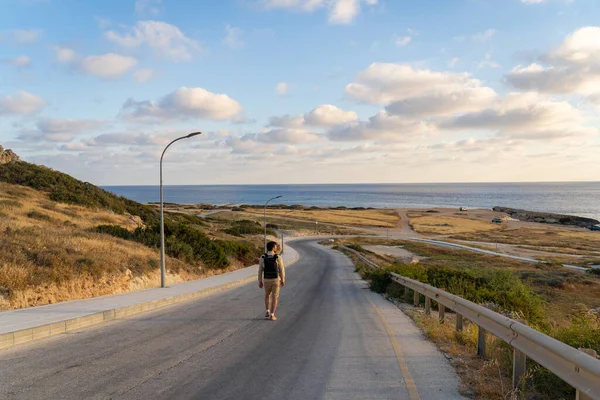 Backpacker hikes on the island of Cyprus, man with backpack walks along the highway on hill near Mediterranean Sea in Agios Georgios Pegeias. Person walking on coastal road on sunset. Adventure — ストック写真