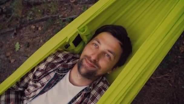 Satisfied Caucasian man lies in green hammock in the forest and looks at camera smiling, top view close-up. Male resting. enjoying nature on hammock outdoors. People relaxation travel concept — Vídeo de Stock