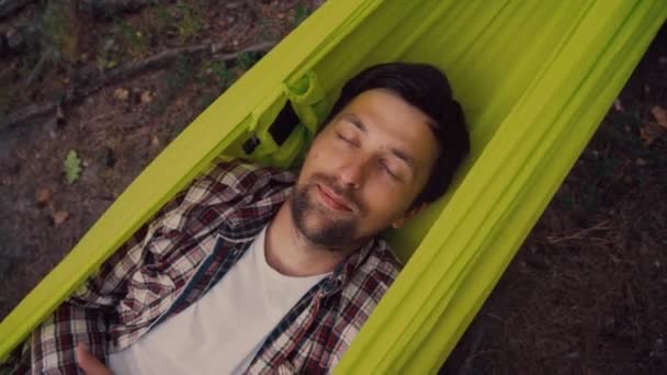 Happy young traveler is resting lying in green hammock with closed eyes and smiling in forest after hiking top view close-up. Outdoors relaxation. Adventurer relaxes in hammock in a coniferous grove — Vídeo de Stock
