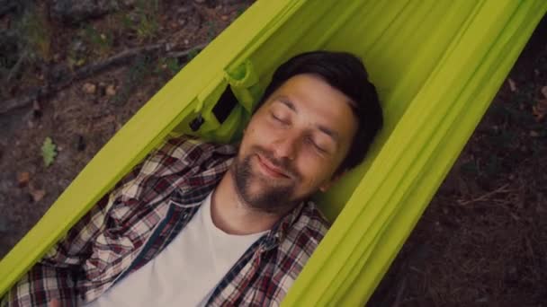 Naturaleza viaje y vacaciones tema. Primer plano del joven caucásico feliz descansando con los ojos cerrados y sonriendo en hamaca verde en el bosque después de caminar en el camping. Gente relajación viaje concepto — Vídeo de stock