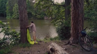 Male cyclist attaches hammock strap to tree in clearing in forest by lake on bicycle trip. Traveler on bike sets green hammock between trees in camping outdoors. Preparing place to rest after cycling