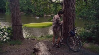 Male cyclist attaches hammock strap to tree in clearing in forest by lake on bicycle trip. Traveler on bike sets green hammock between trees in camping outdoors. Preparing place to rest after cycling