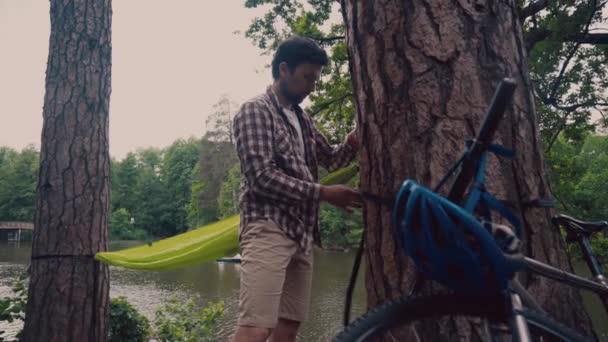 Male tourist sets hammock arrived at camping place near lake on bicycle. Man cyclist setting up hammock in forest by river. Traveler tying hammock in park by pond. Guy attaches hammock strap to tree — Wideo stockowe