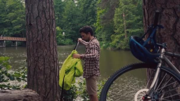 男子观光台吊床骑自行车到达湖边的野营处。骑自行车的人在河边的森林里架起吊床.旅行者把吊床绑在一个接一个的池塘里.那家伙把吊袜带绑在树上 — 图库视频影像