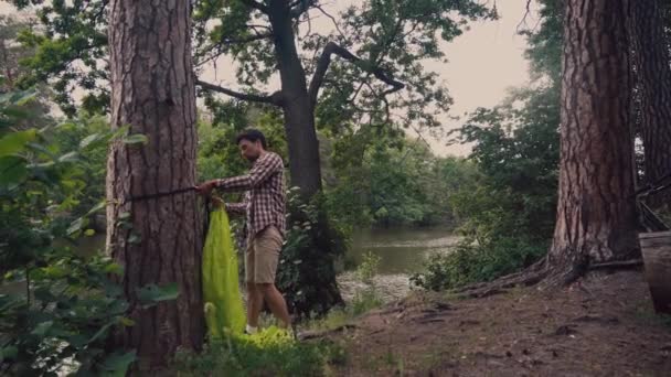 Cykelturist anländer till gläntan i skogen vid sjön och förbereder campingplatser genom att hänga grön hängmatta mellan träd med utsikt över sjön. Man sätter upp hängmatta på cykel resa vid floden — Stockvideo