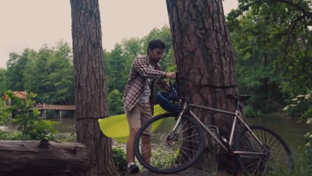 Bicycle tourist arrives at clearing in forest by lake and prepares camping sites by hanging green hammock between trees overlooking lake. Man sets up hammock on bike trip by the river — Video