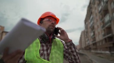 Frustrated construction foreman talking with an employee on his cell phone. Angry supervisor with papers, unhappy with deadline for completion of work, swears from stress while talking on phone
