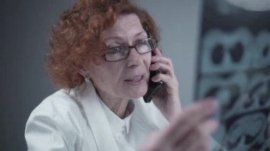Elderly caucasian woman doctor analyze x-ray image and talk with mobile phone about diagnostics in medical office. Doctor specialist working with CT scan, talk to cellphone about radiography results