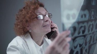 Happy adult woman doctor radiologist discussing CT scan of patients lungs with colleagues by phone in hospital office. Pulmonologist examines MRI or pulmonography and uses a cell phone. Chest x-ray