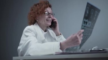Elderly caucasian woman doctor analyze x-ray image and talk with mobile phone about diagnostics in medical office. Doctor specialist working with CT scan, talk to cellphone about radiography results