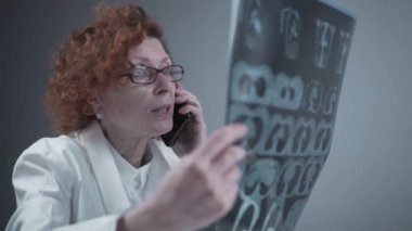 Happy adult woman doctor radiologist discussing CT scan of patients lungs with colleagues by phone in hospital office. Pulmonologist examines MRI or pulmonography and uses a cell phone. Chest x-ray
