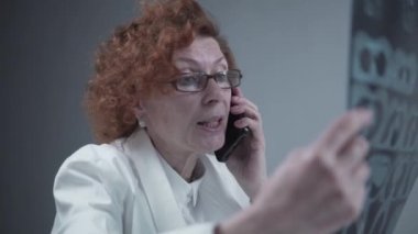 Elderly caucasian woman doctor analyze x-ray image and talk with mobile phone about diagnostics in medical office. Doctor specialist working with CT scan, talk to cellphone about radiography results
