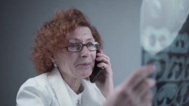 Happy adult woman doctor radiologist discussing CT scan of patients lungs with colleagues by phone in hospital office. Pulmonologist examines MRI or pulmonography and uses a cell phone. Chest x-ray