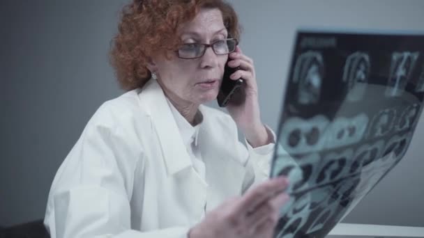 Anciana caucásica mujer médico analizar la imagen de rayos X y hablar con el teléfono móvil acerca de los diagnósticos en el consultorio médico. Médico especialista que trabaja con la tomografía computarizada, hable con el teléfono celular sobre los resultados de la radiografía — Vídeo de stock