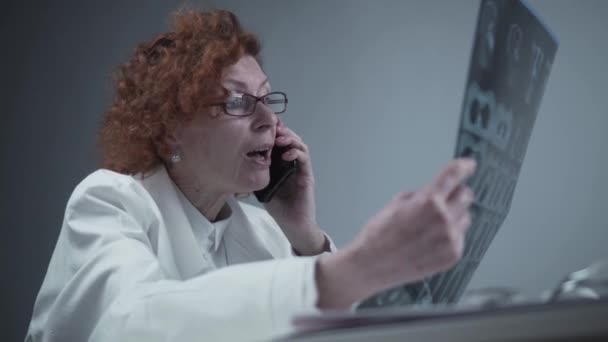 Doctora mirando la tomografía computarizada hablando con su colega en el teléfono inteligente en la oficina del hospital. Doctor al teléfono en la clínica, examinando tomografías computarizadas y discutiendo sus hallazgos. Radiólogo examen de rayos X de pulmón — Vídeo de stock