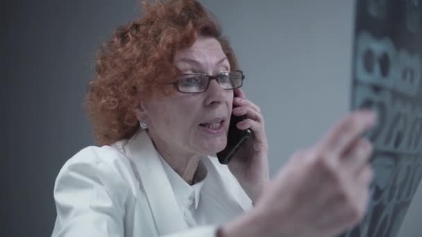 Elderly caucasian woman doctor analyze x-ray image and talk with mobile phone about diagnostics in medical office. Doctor specialist working with CT scan, talk to cellphone about radiography results — Stok video