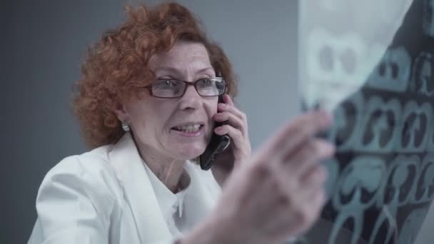 Женщина-врач смотрит на рентген, разговаривает с коллегой по смартфону в больничном кабинете. Доктор по телефону в клинике, изучает компьютерную томографию и обсуждает свои выводы. Рентгенологическое исследование легких — стоковое видео
