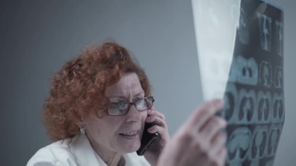 Anciana caucásica mujer médico analizar la imagen de rayos X y hablar con el teléfono móvil acerca de los diagnósticos en el consultorio médico. Médico especialista que trabaja con la tomografía computarizada, hable con el teléfono celular sobre los resultados de la radiografía — Vídeo de stock